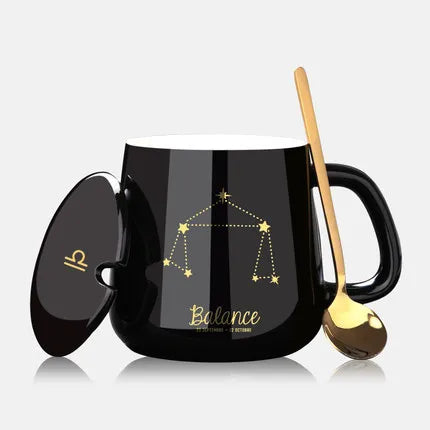 Black mug with Libra zodiac design, gold spoon, and lid on a light gray background