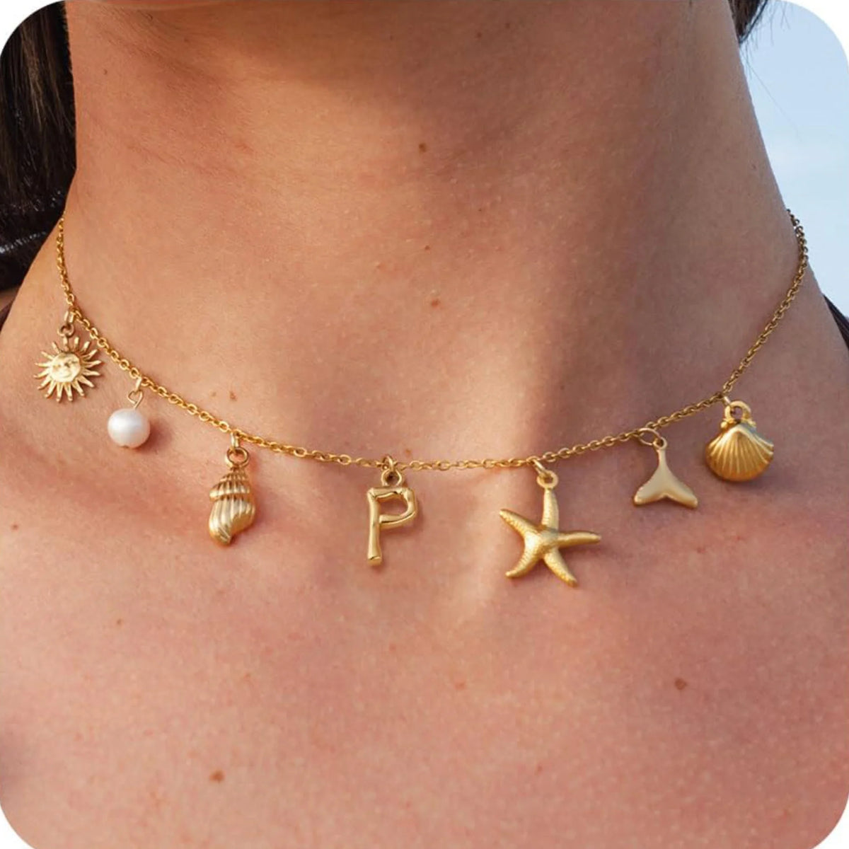 Gold necklace with various charms including a sun, shell, starfish, and letter 'P' on a person's neck.